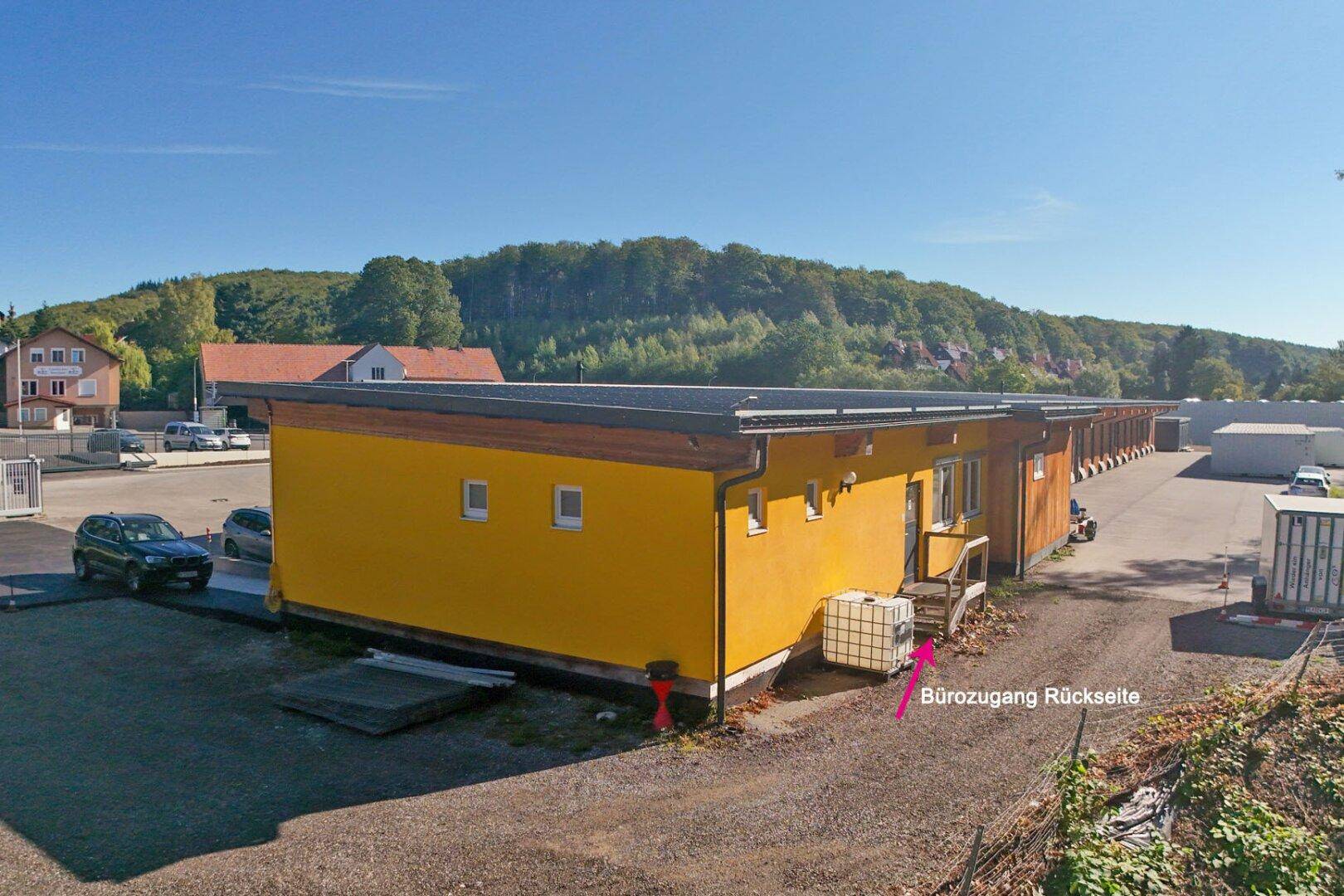 Seitenansicht des Gewerbegebäudes mit gelber Fassade und einem weiteren Zugangsbereich im Grünen.