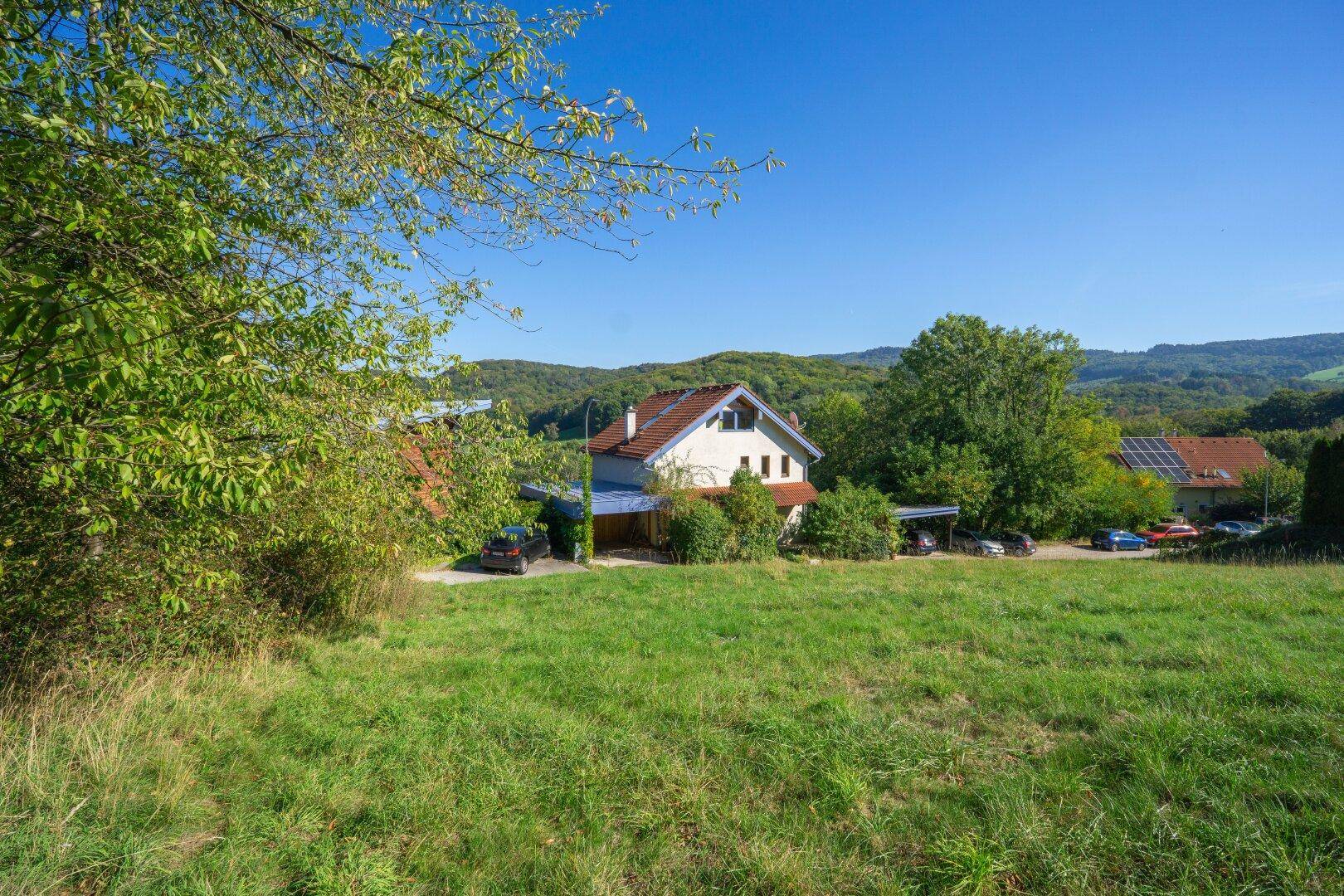 Weitläufige grüne Landschaft mit Wohnhäusern und bewaldeten Hügeln unter klarem Himmel.