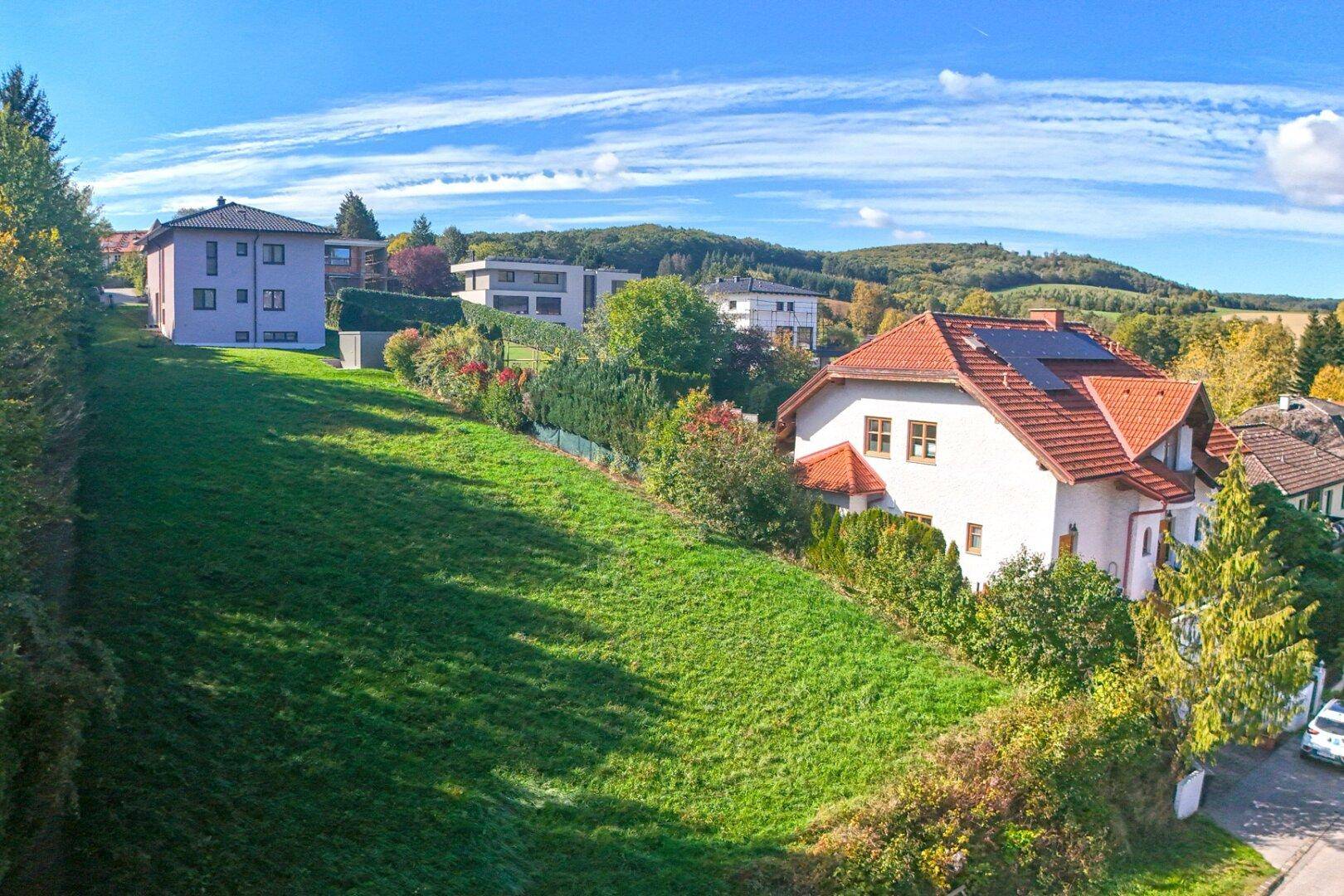 Weitwinkelaufnahme des abfallenden Grundstücks mit angrenzenden Häusern und einem weiten Blick über die grüne Landschaft.