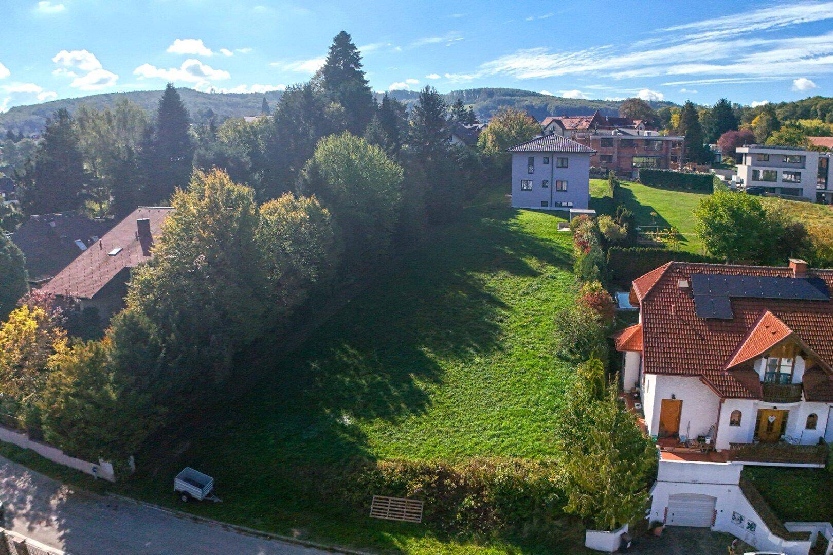 Luftaufnahme eines großen, sonnigen Grundstücks umgeben von Bäumen und Wohnhäusern in hügeliger Landschaft.