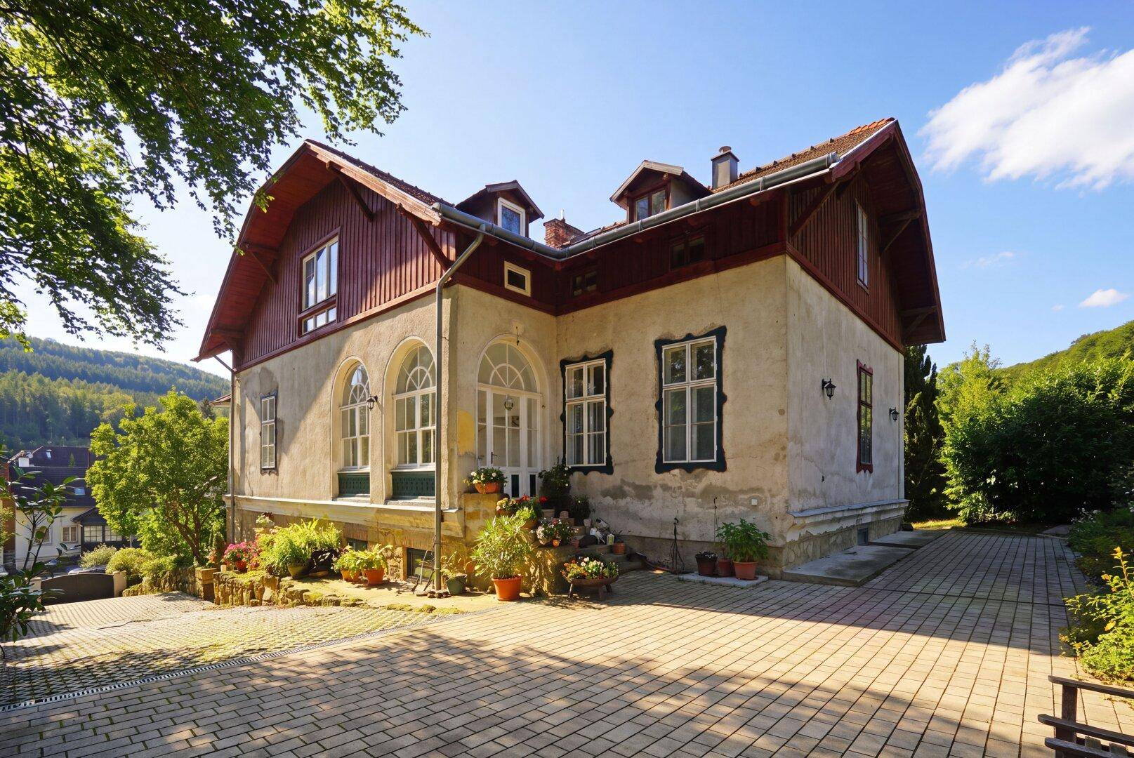 Historisches Gebäude mit rotem Dach, gepflastertem Vorplatz und umgeben von viel Grün.