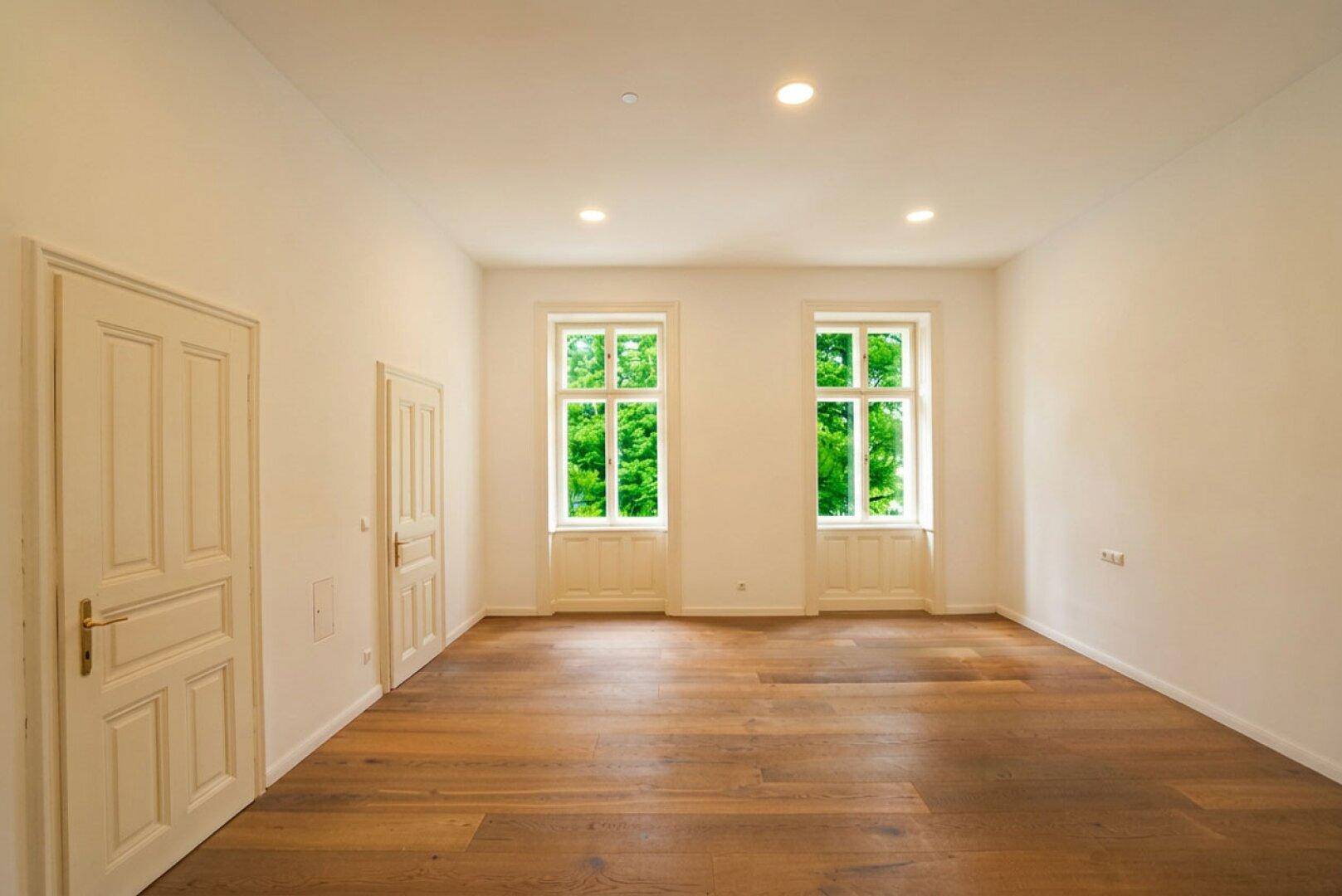 Helles, leeres Zimmer mit Holzboden und zwei Fenstern, die einen Blick ins Grüne bieten.