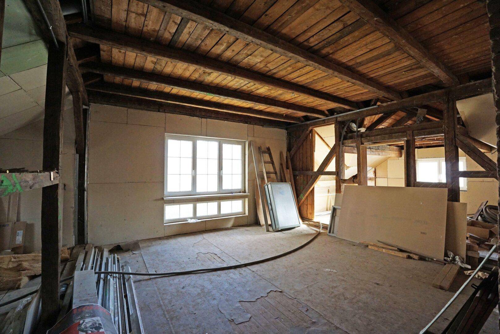 Unausgebauter Dachboden mit sichtbaren Holzbalken, Fenster und Potenzial für weiteren Ausbau.
