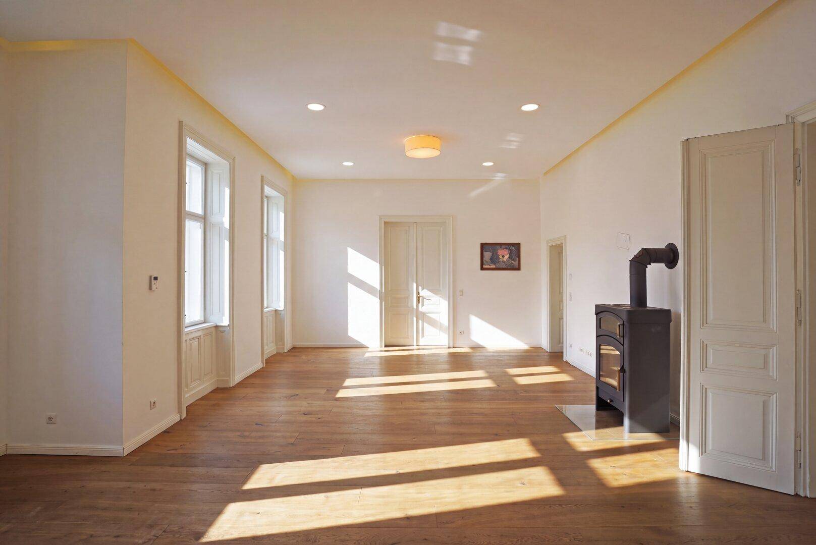 Geräumiges Wohnzimmer mit Holzboden, Kaminofen und hohen Decken, durchflutet von natürlichem Licht.