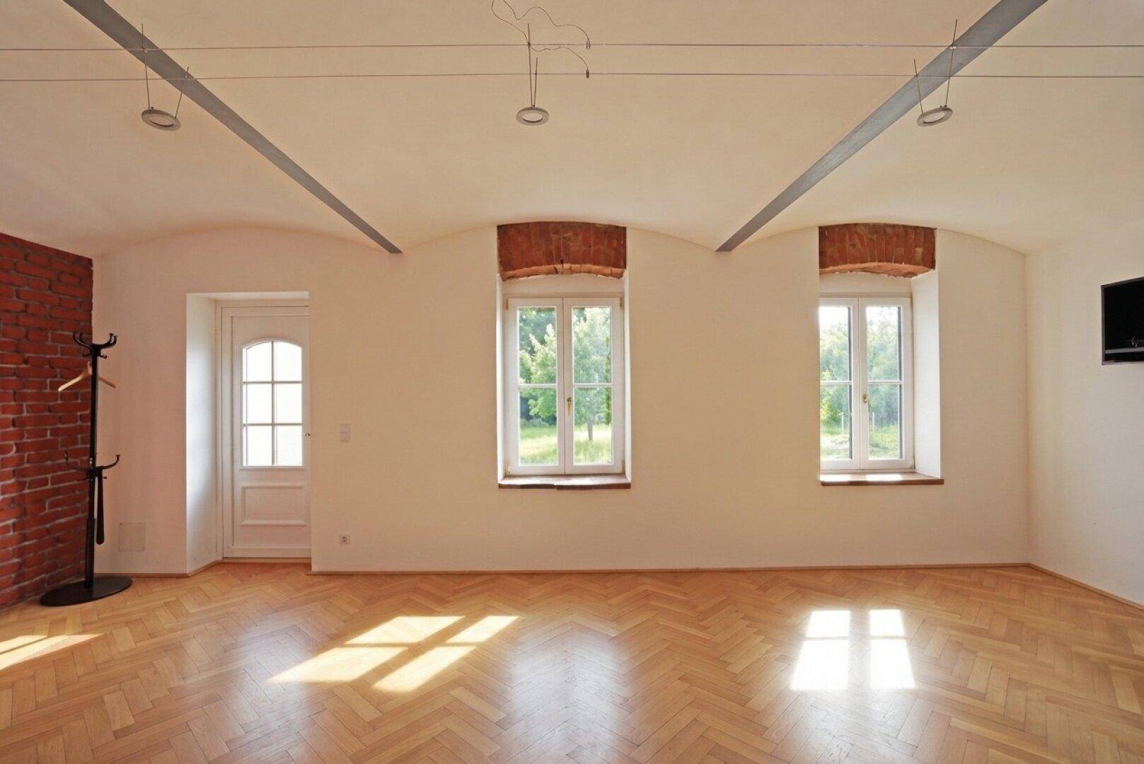 Geräumiger Raum mit Fischgrätparkett, freiliegender Ziegelwand und hohen Decken.