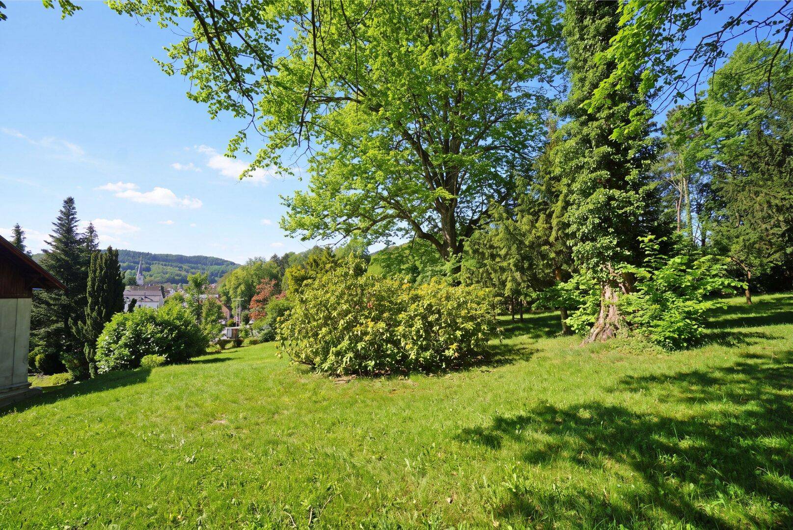 Weitläufiger grüner Garten mit altem Baumbestand und Blick auf die umliegende Landschaft.
