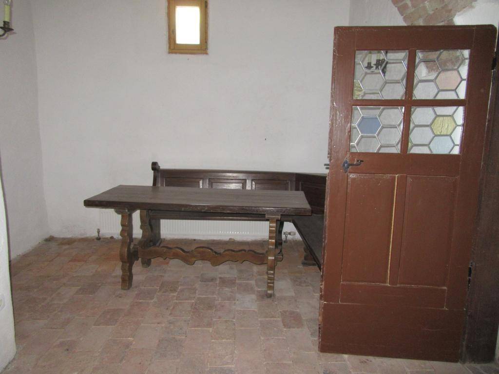 Rustikaler Raum mit Terrakotta-Bodenfliesen, einem Holztisch und einer Bank, mit kleiner Fensteröffnung.