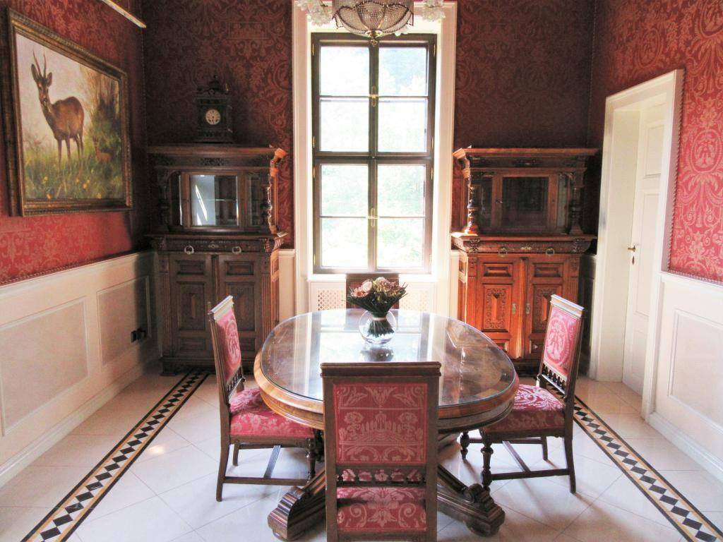 Elegantes Esszimmer mit rundem Tisch, Stühlen und gemusterten roten Tapeten, viel Tageslicht.