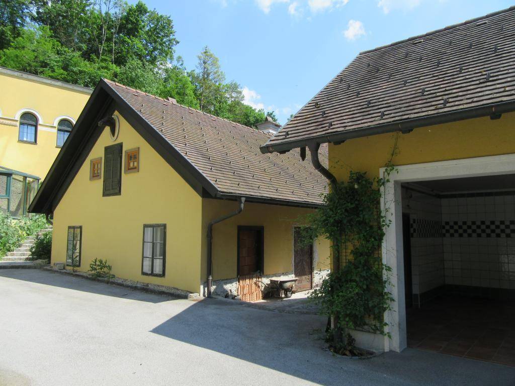 Gelbes Nebengebäude mit Ziegeldach und offenem Garagenbereich, umgeben von Grün.