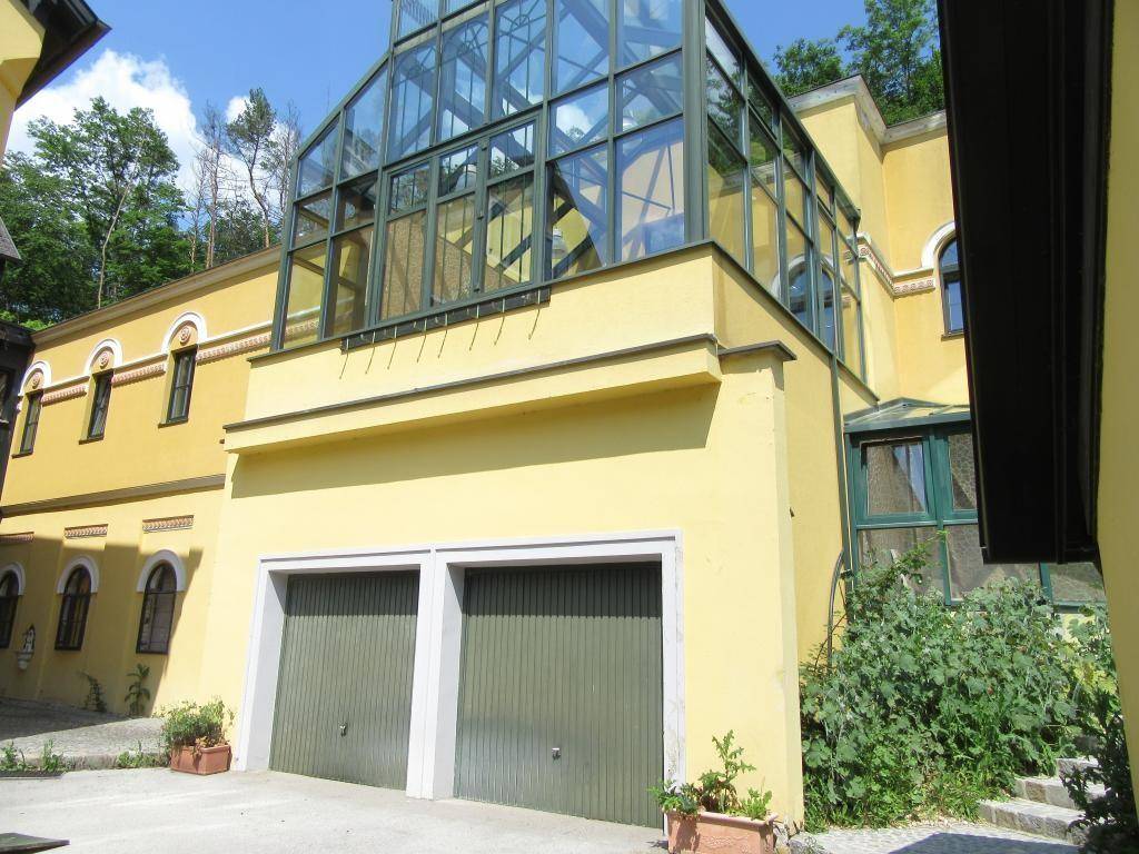 Gelbes Gebäude mit großem verglastem Wintergarten und zwei Garagentoren, umgeben von Pflanzen.