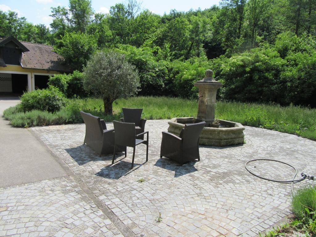 Gepflasterter Außenbereich mit Sitzgelegenheiten und einem steinernen Brunnen, umgeben von Grün.