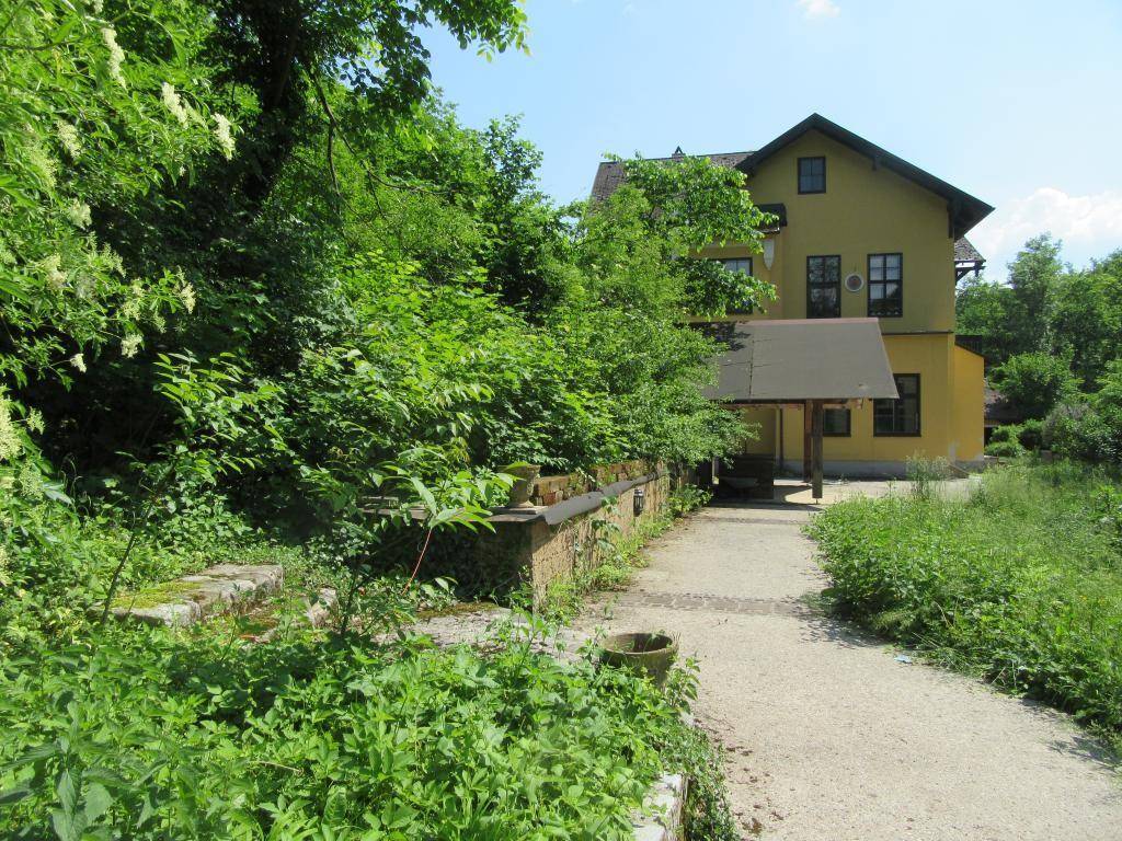 Seitenansicht eines gelben Gebäudes, umgeben von üppiger grüner Vegetation und einem Gartenweg.