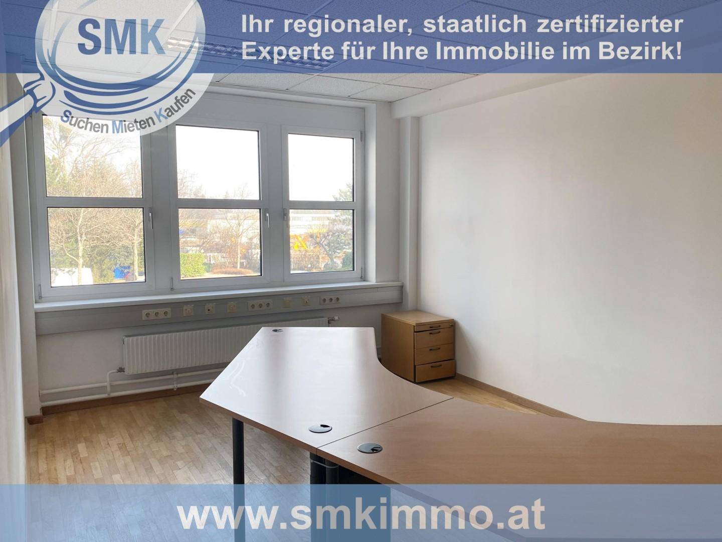 Leeres Eckbüro mit Schreibtisch und kleinem Rollcontainer, großen Fenstern und hellem Holzfußboden.