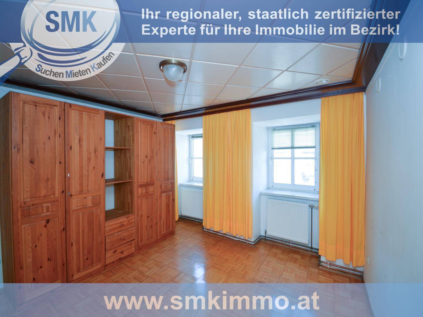 Schlafzimmer mit großen Holzschränken, Parkettboden und zwei Fenstern mit gelben Vorhängen.