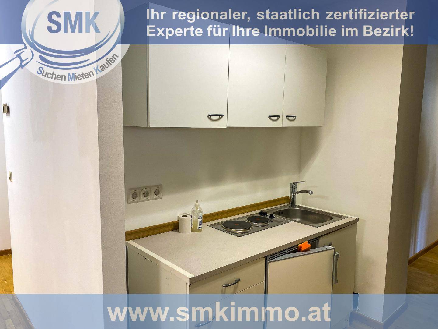Kleine Küchenzeile mit Spüle, zwei Kochplatten, einem Kühlschrank und weißen Hängeschränken.