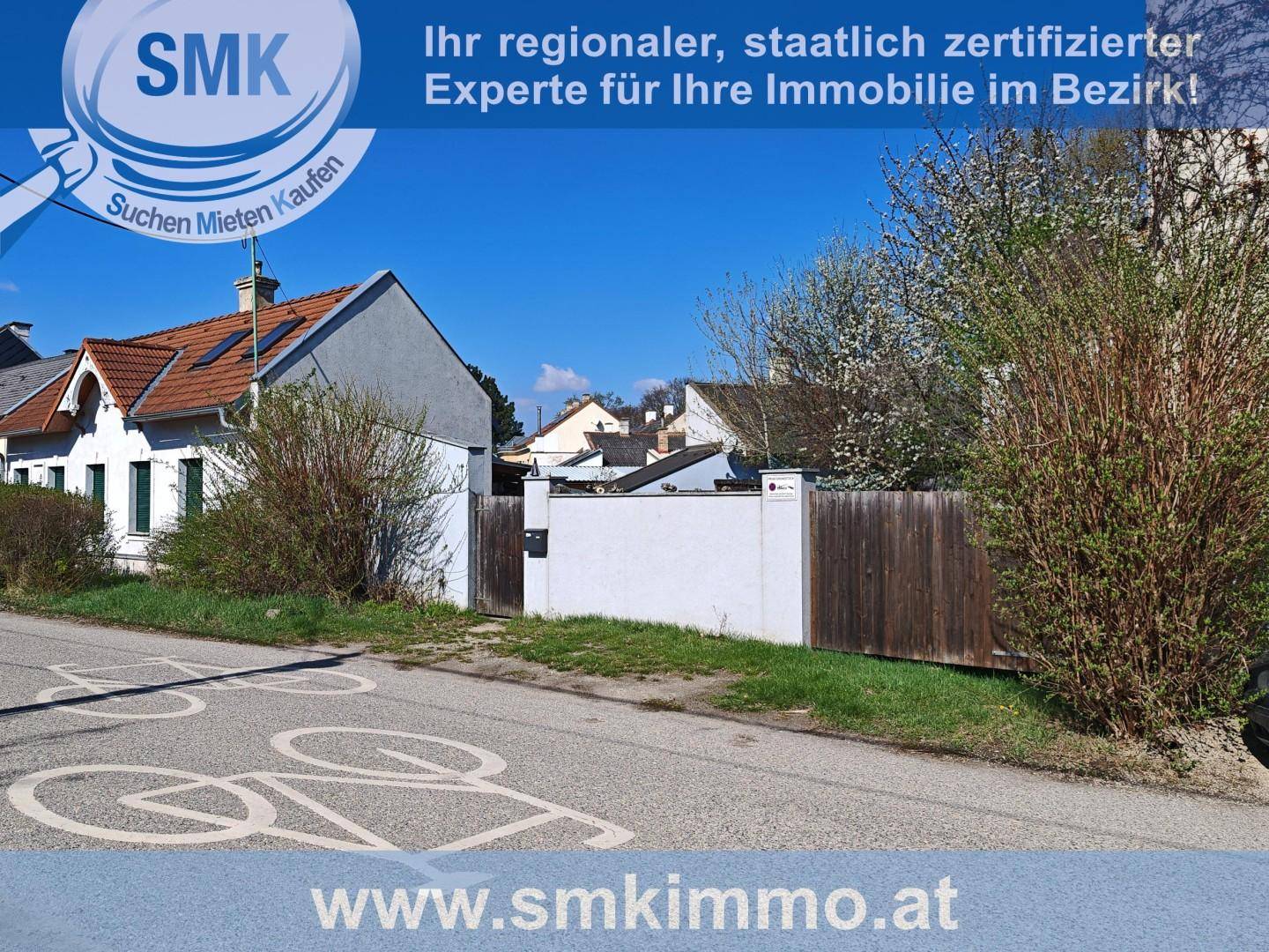 Blick auf ein Haus mit Zaun und Straße, umgeben von Bäumen und Sträuchern in ländlicher Gegend.