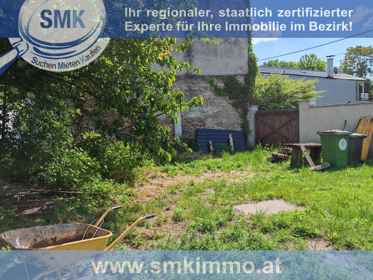 Verwilderter Gartenbereich mit Gras, Sträuchern, einer Schubkarre und alter Ziegelmauer.