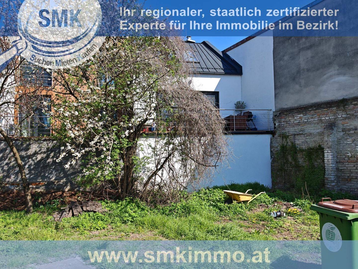 Gartenbereich mit einem blühenden Baum, einer Schubkarre und einem Gebäude mit Balkon im Hintergrund.