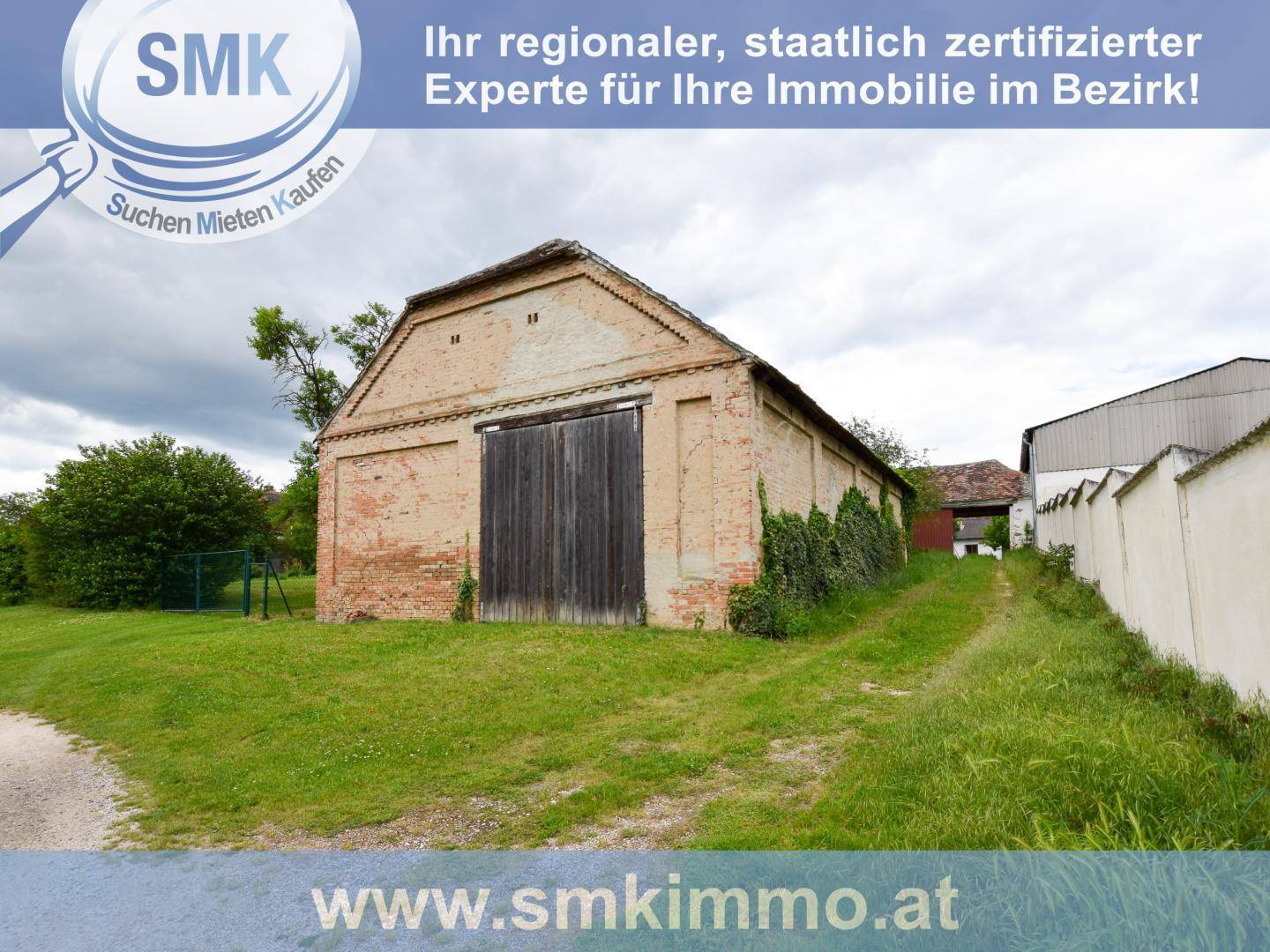 Große alte Backsteinscheune mit Holztor und weitläufiger Grünfläche davor unter bewölktem Himmel.