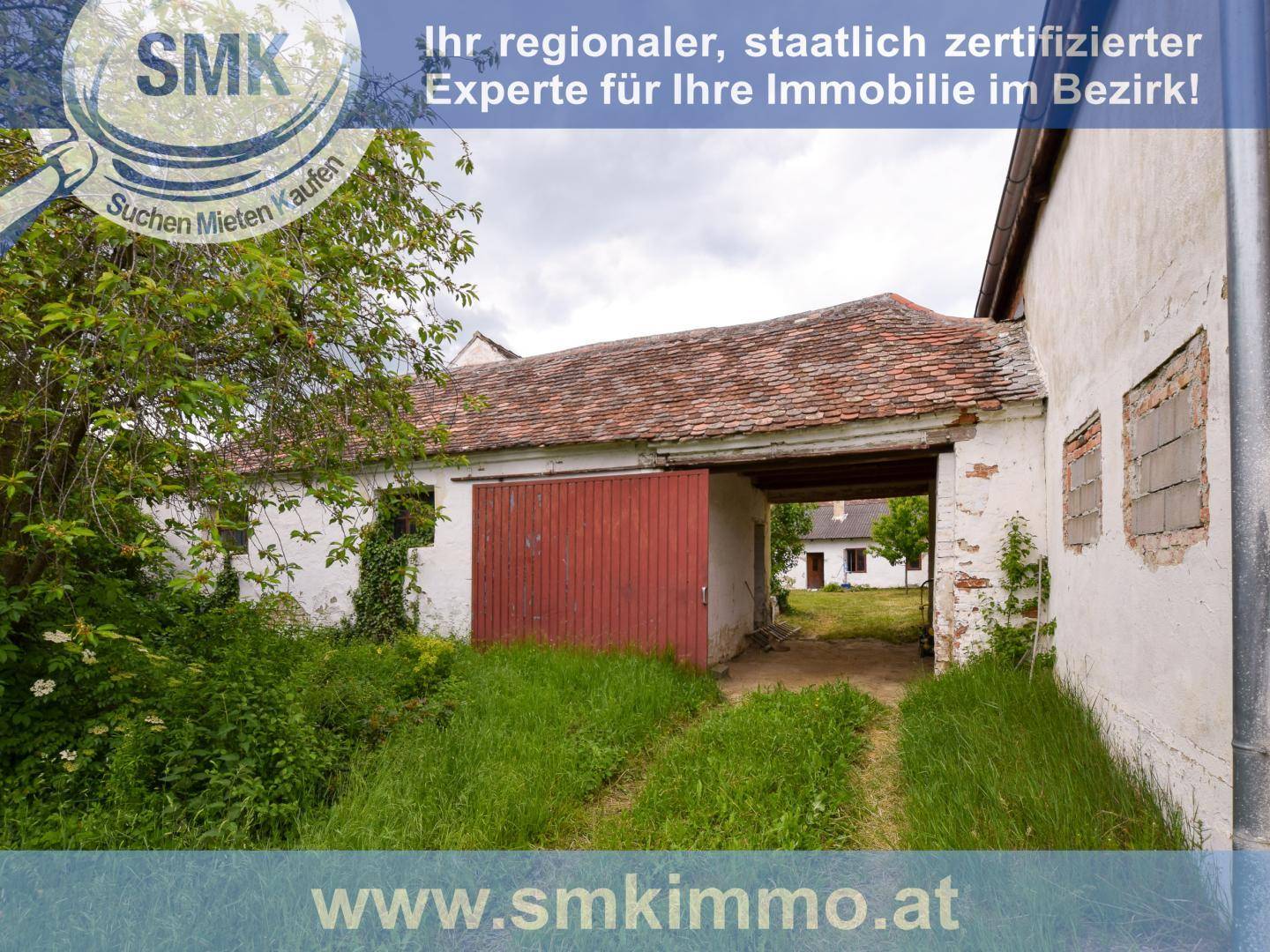 Altes weißes Gebäude mit großem roten Holztor und überwucherter Vegetation am Eingang.