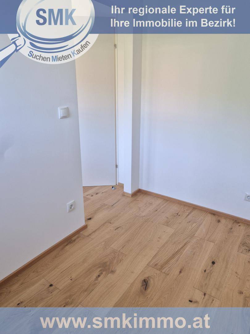 Ecke eines hellen Zimmers mit Holzboden und weißen Wänden, eine Tür ist sichtbar.