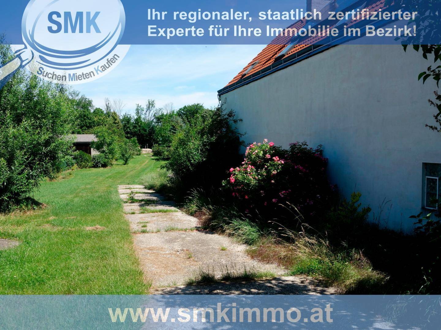 Seitenansicht des Hauses mit grünem Gartenweg und blühenden Sträuchern unter blauem Himmel.