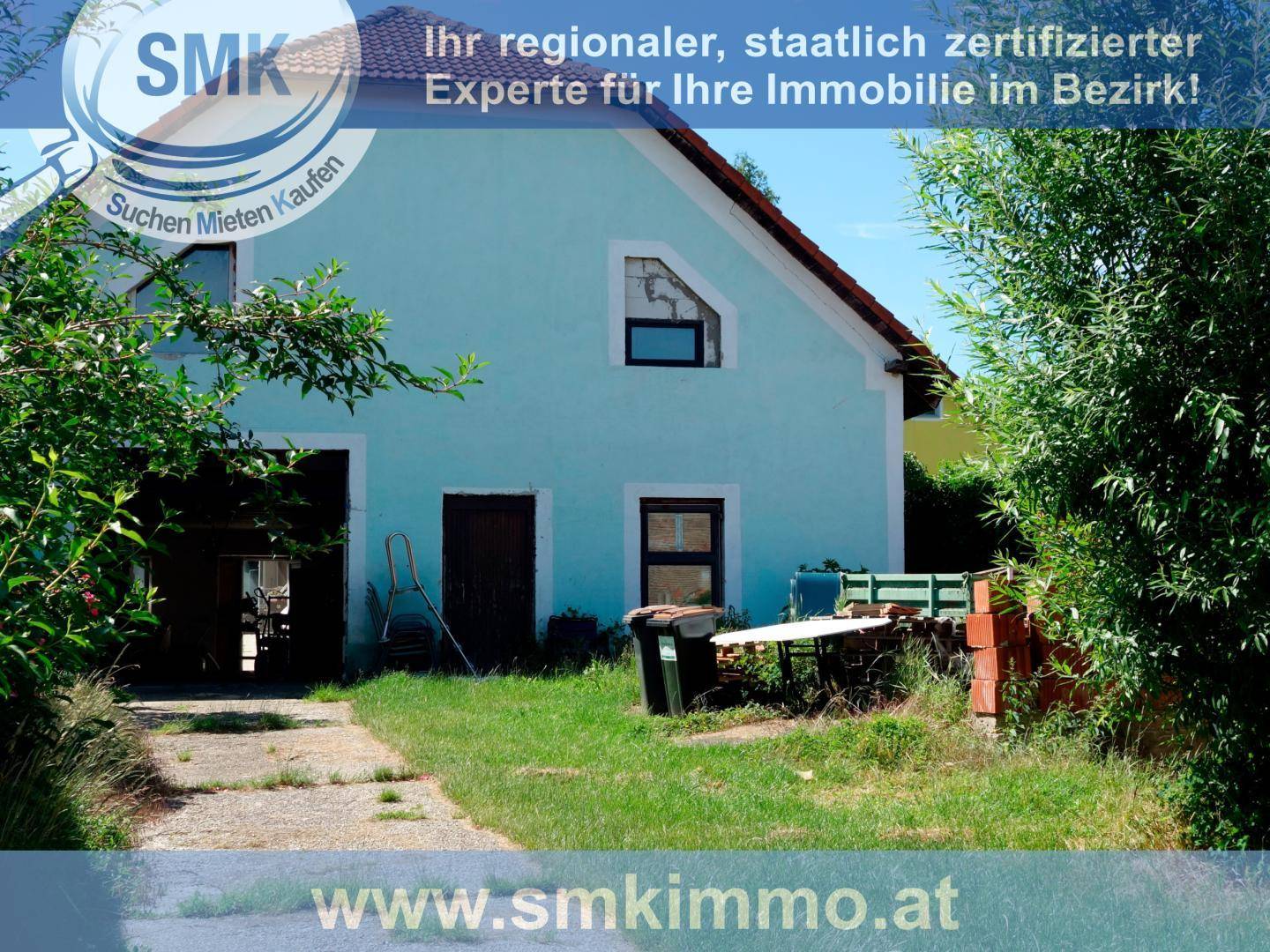 Rückseite eines hellblauen Hauses mit offenem Garagenbereich und Garten, renovierungsbedürftig.