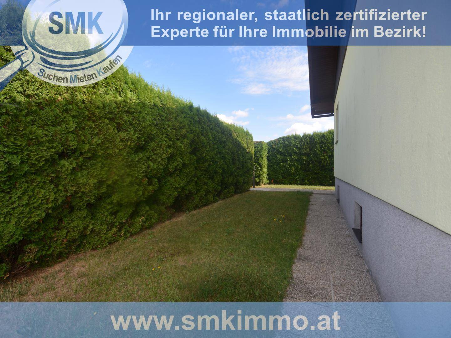 Seitenansicht des Hauses mit schmalem Gehweg, Rasenfläche und hoher Hecke unter blauem Himmel.