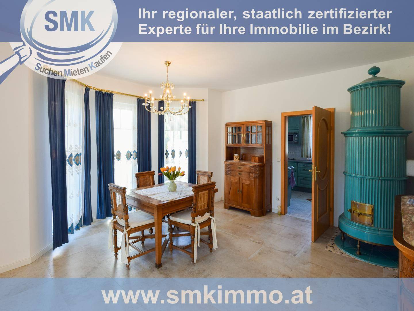 Gemütlicher Essbereich mit traditionellem Kachelofen, Holztisch und blauen Vorhängen.