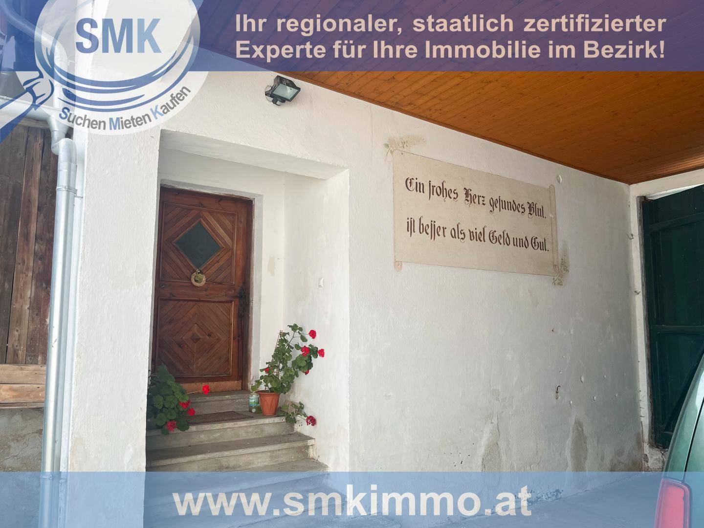 Überdachter Eingangsbereich mit einer Holztür und einer Inschrift an der weiß getünchten Wand.