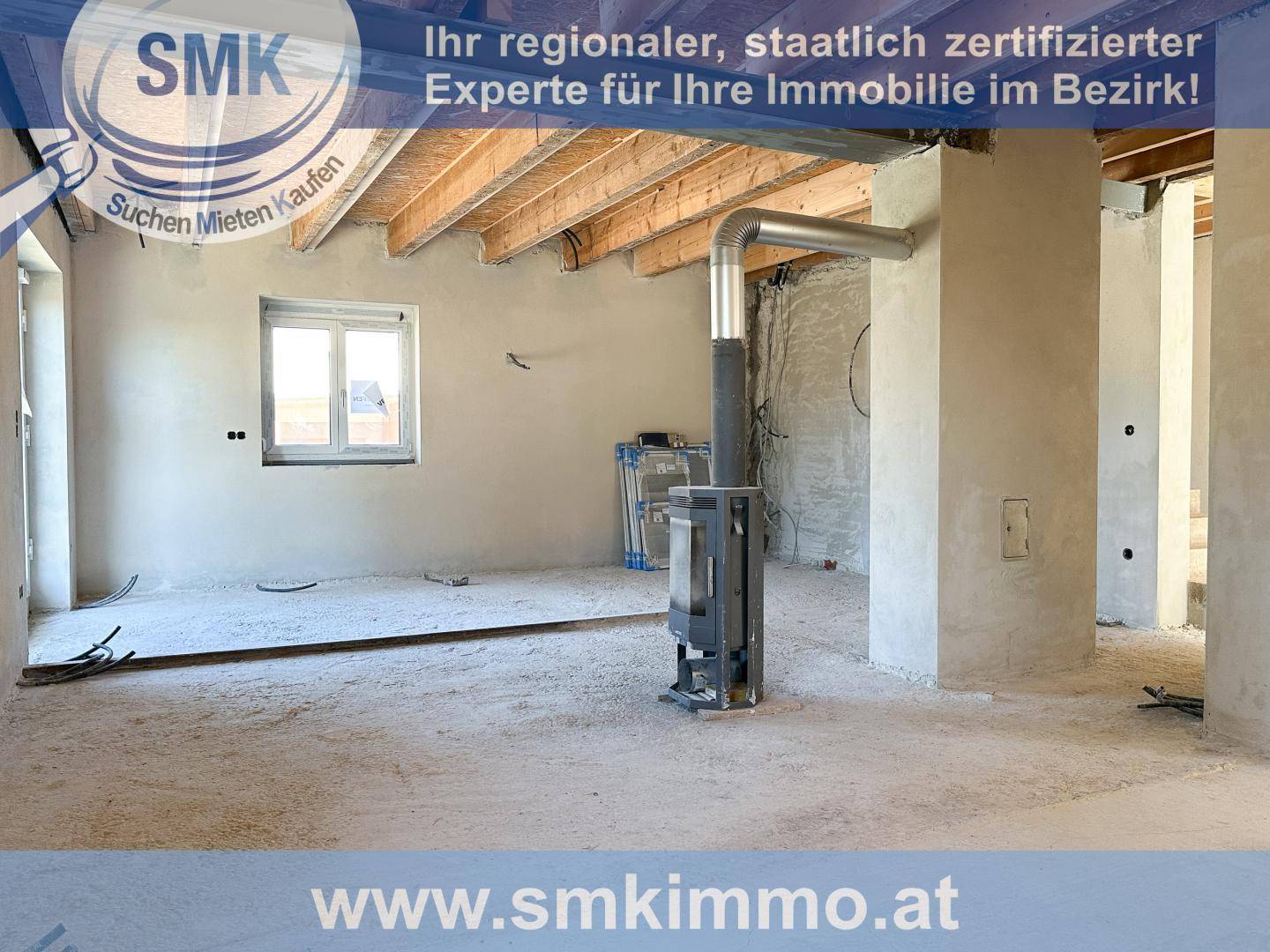 Unfertiger Wohnraum mit Holzofen, sichtbaren Deckenbalken und einem Fenster.