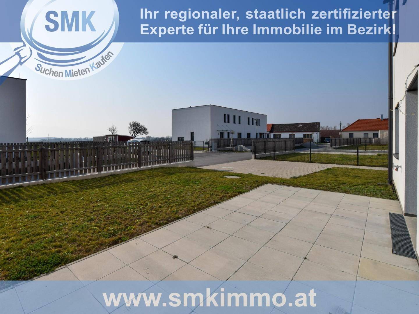 Außenbereich mit gepflasterter Terrasse und Rasenfläche, umgeben von einem Zaun und weiteren Gebäuden.