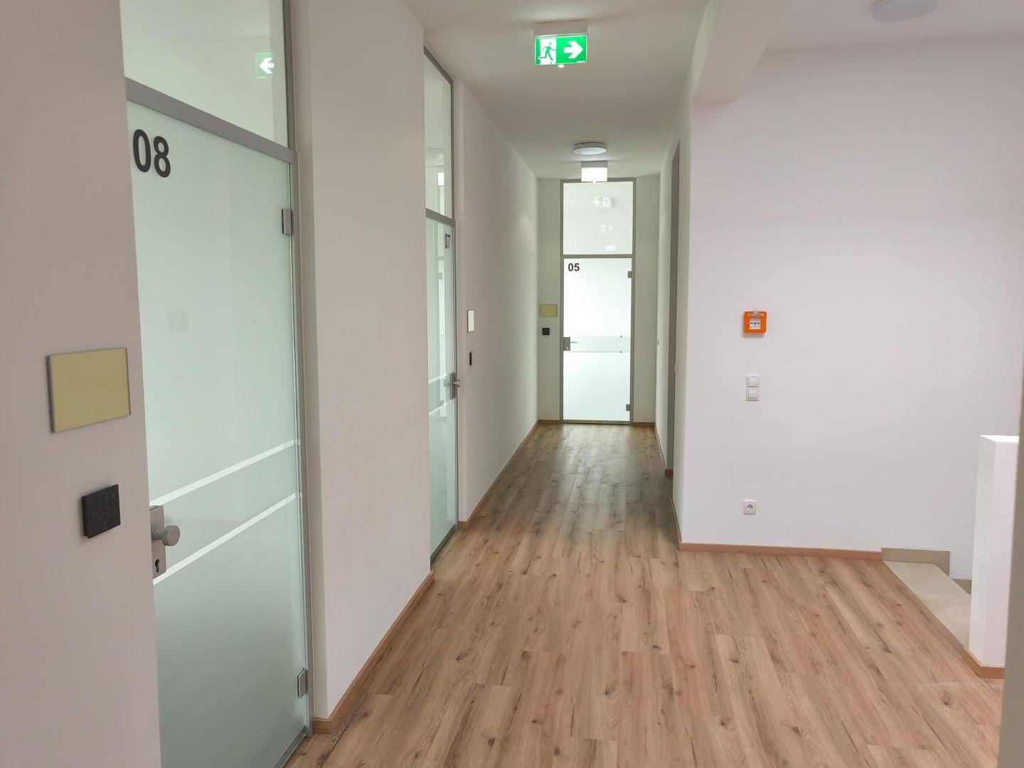 Moderner Büroflur mit Laminatboden, weißen Wänden und Glastüren zu den einzelnen Büros.