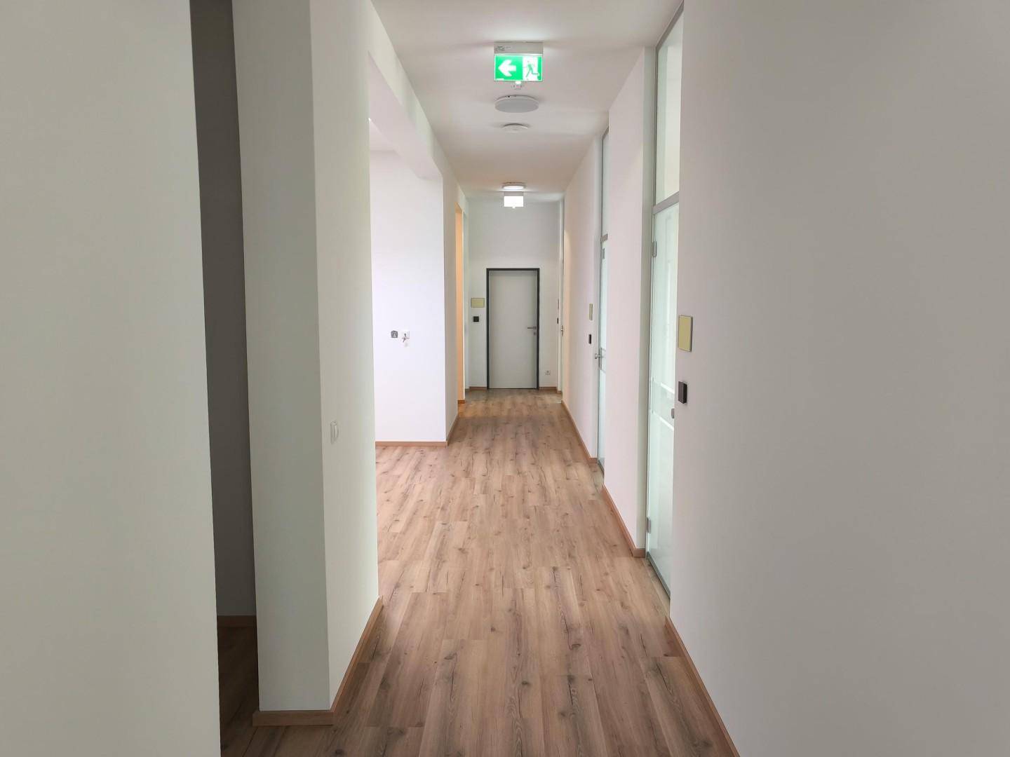 Langer, heller Büroflur mit Laminatboden und weißen Wänden, der zu weiteren Räumen führt.