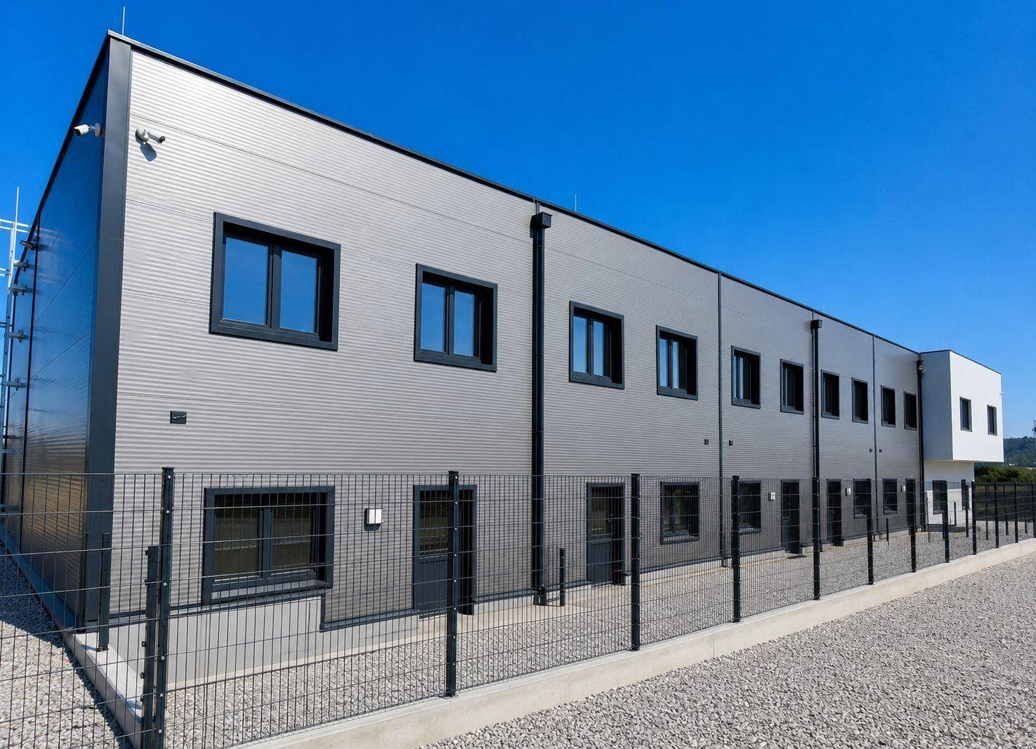 Seitenansicht eines modernen Gewerbegebäudes mit Fenstern, Sicherheitszaun und Kameraüberwachung.