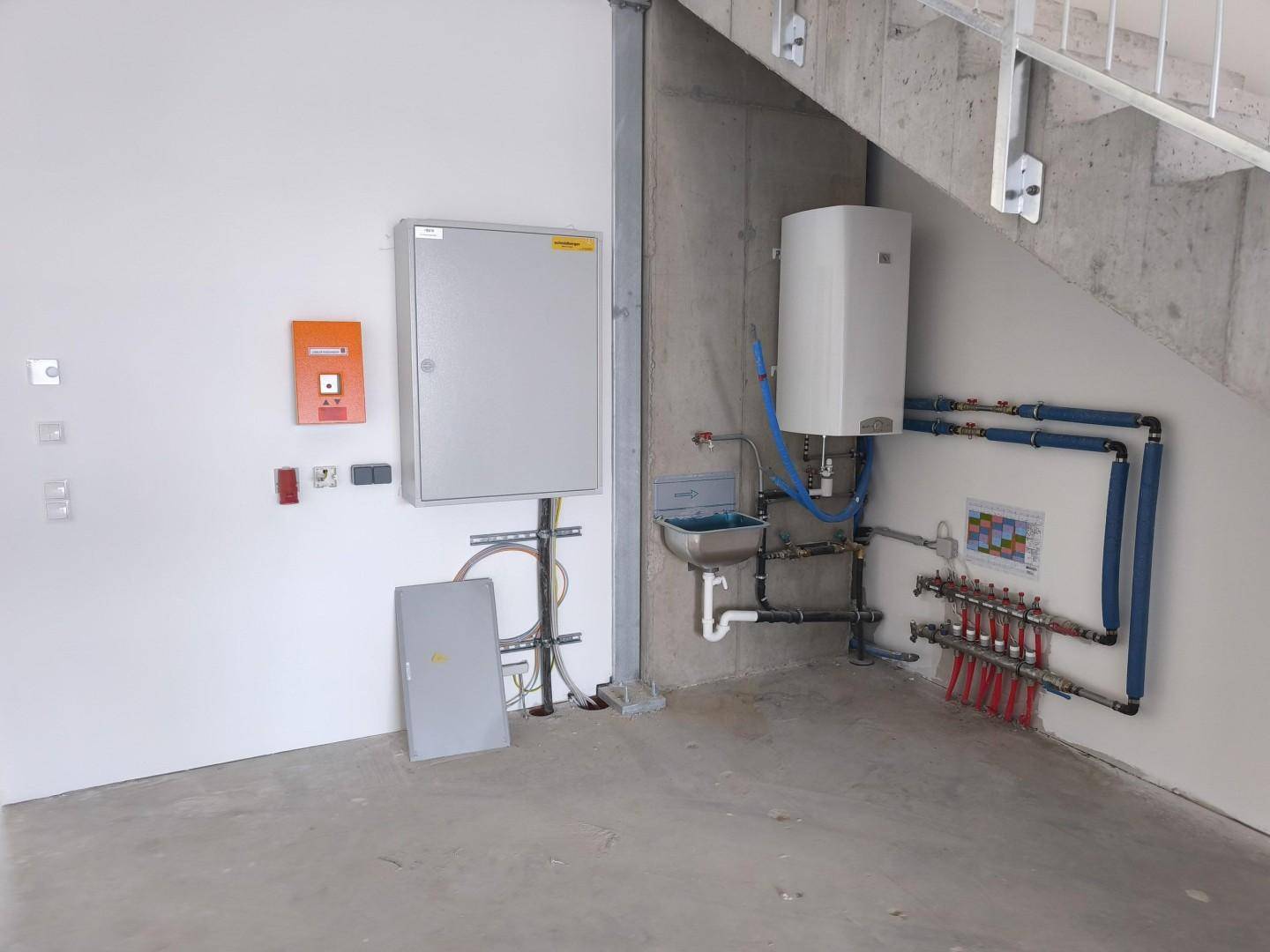 Technikbereich unter der Treppe mit Wasserleitungen, Boiler und Elektroschrank in einem Neubau.