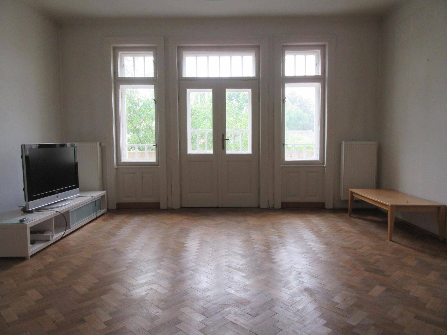 Heller, leerer Raum mit Fischgrätparkett und großen Fenstern, die viel Tageslicht hereinlassen.