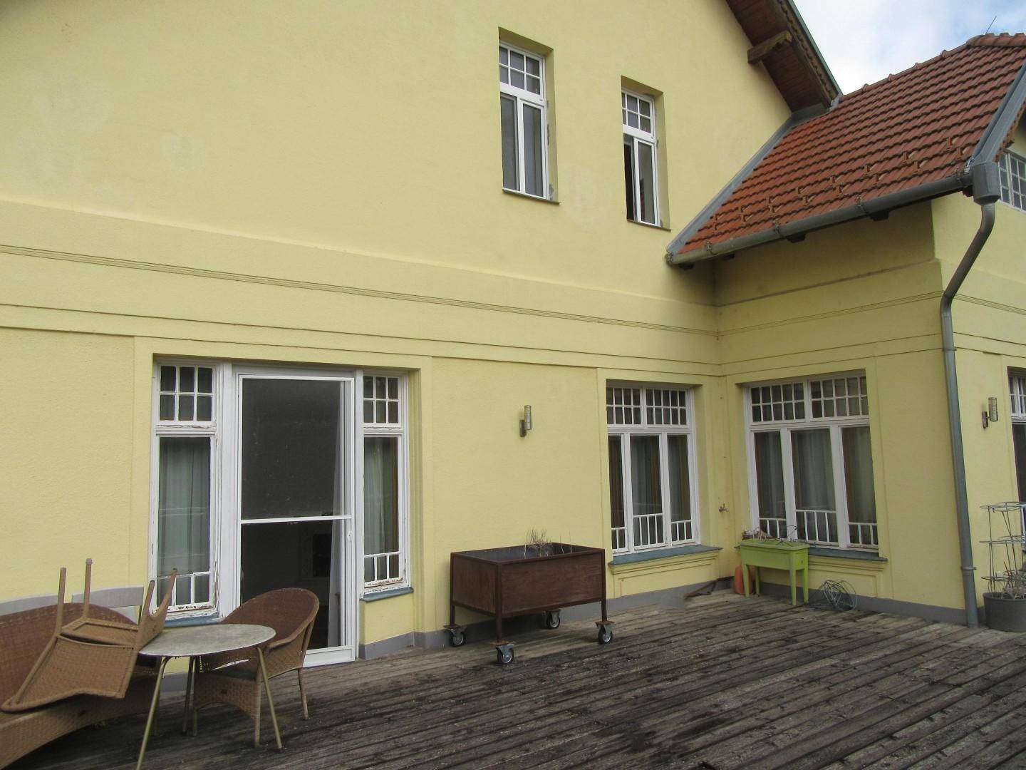 Gelbe Hausfassade mit großer Holzterrasse und Gartenmöbeln, umgeben von Grün.