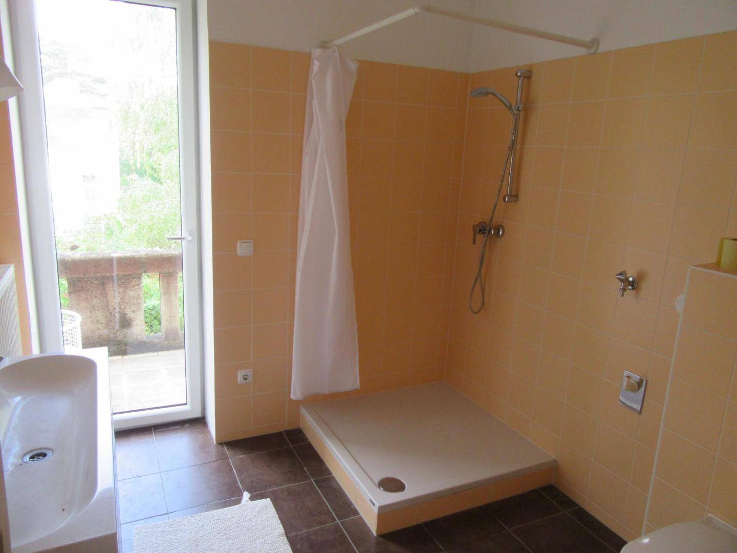 Badezimmer mit Duschbereich, Waschbecken und Zugang zu einem Balkon, hell gefliest.