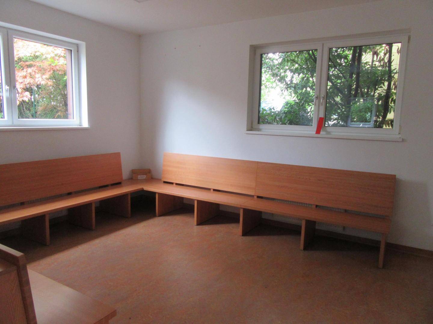 Raum mit Holzbänken und großen Fenstern, die einen Blick ins Grüne bieten.
