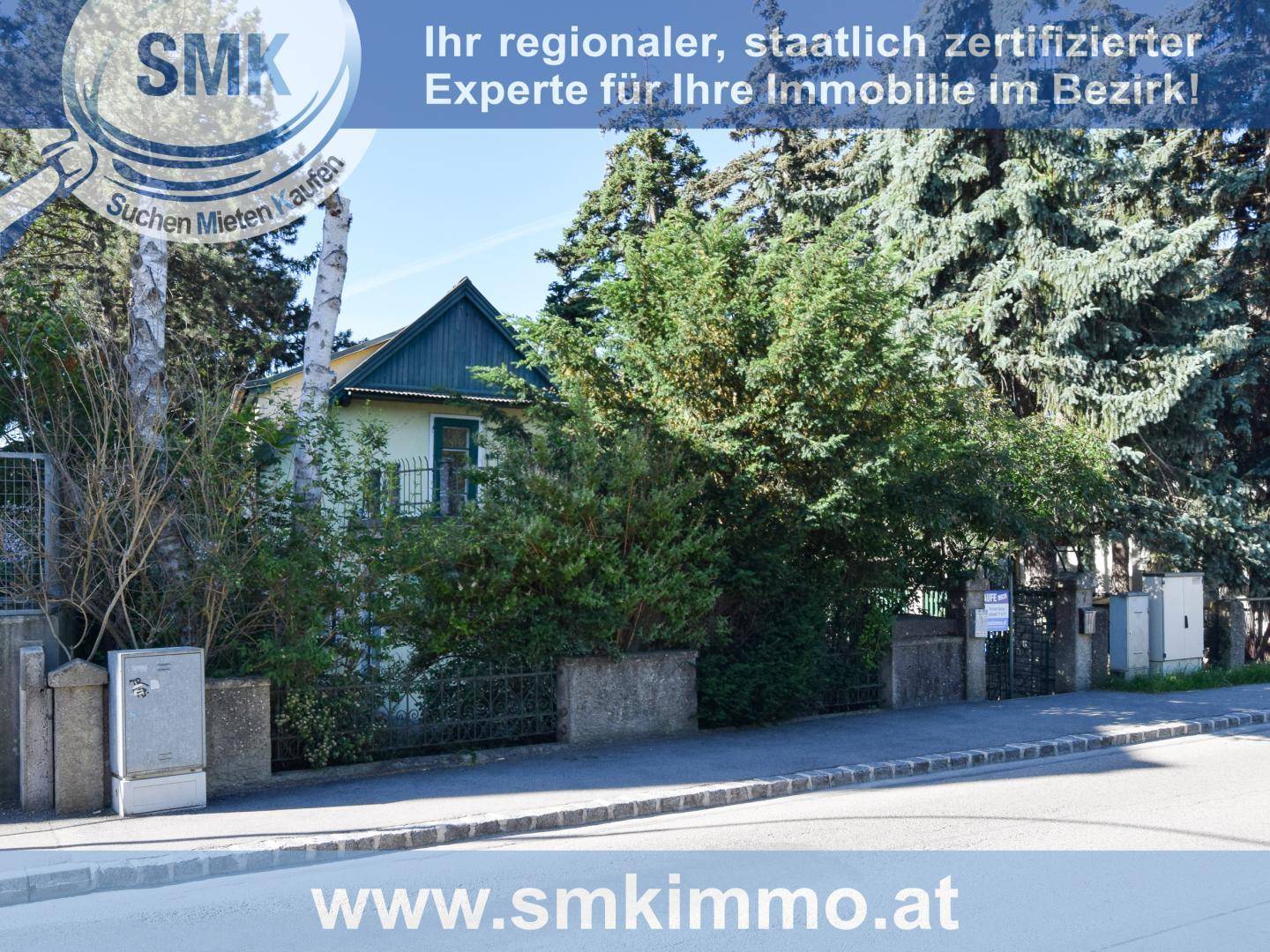 Fassade eines Hauses, umgeben von dichtem Grün, mit Gehweg und Straße im Vordergrund der Immobilie.