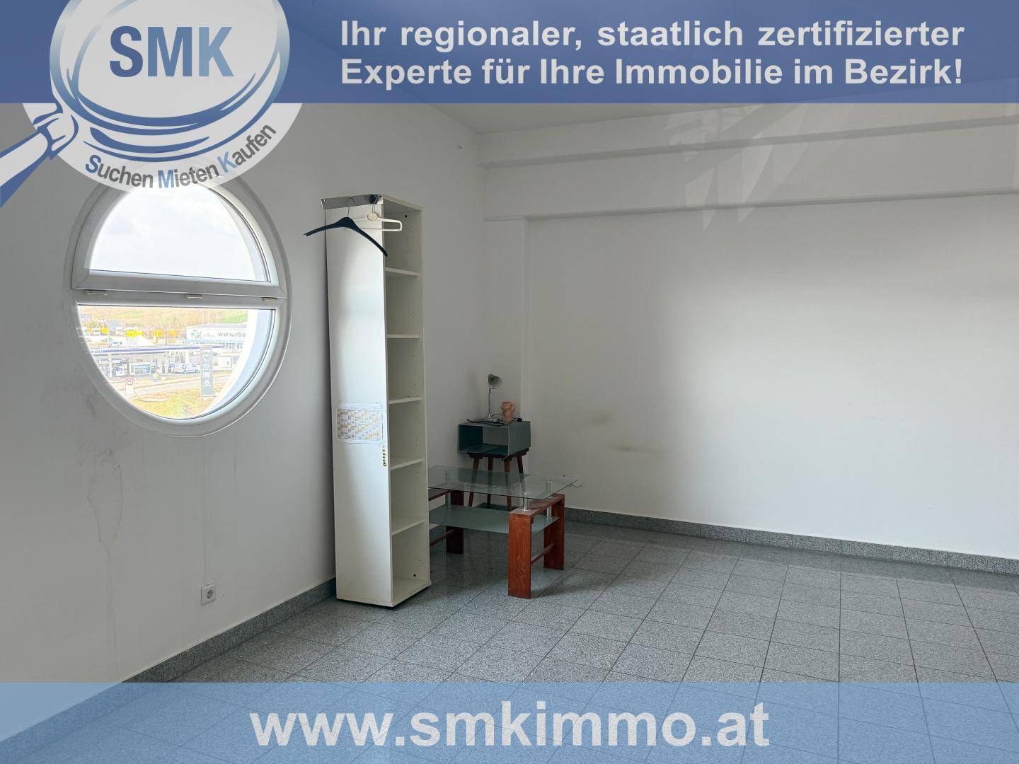 Heller Raum mit Fliesenboden, einem runden Fenster und einfacher Einrichtung, die auf eine Wohnnutzung hindeutet.