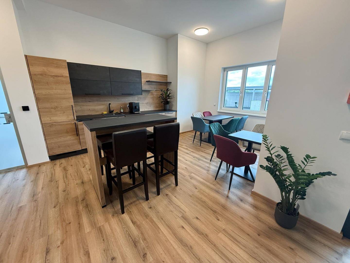 Moderner offener Bereich mit Holzküche, dunkler Arbeitsplatte und Essbereich mit bunten Stühlen und großen Fenstern.