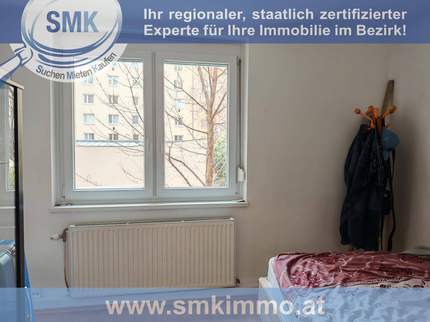 Schlafzimmer mit großem Fenster, Heizkörper und Blick auf umliegende Gebäude und Bäume.