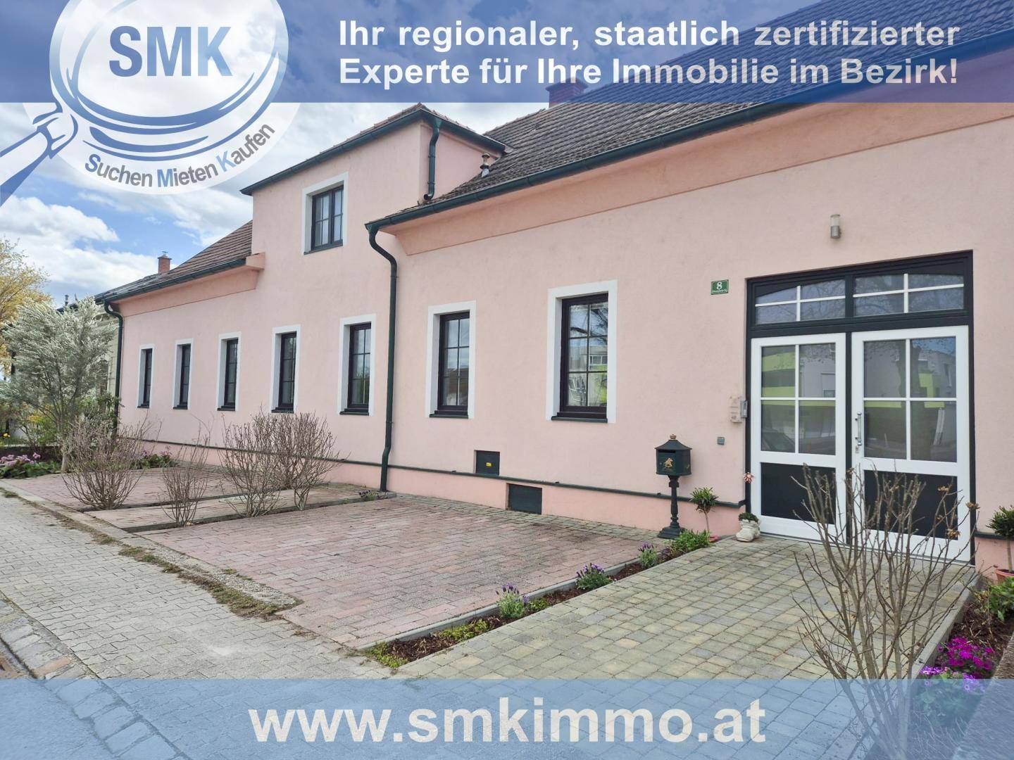 Rosa gestrichenes Haus mit schwarzen Fenstern und Eingangstür, umgeben von einem gepflasterten Bereich.