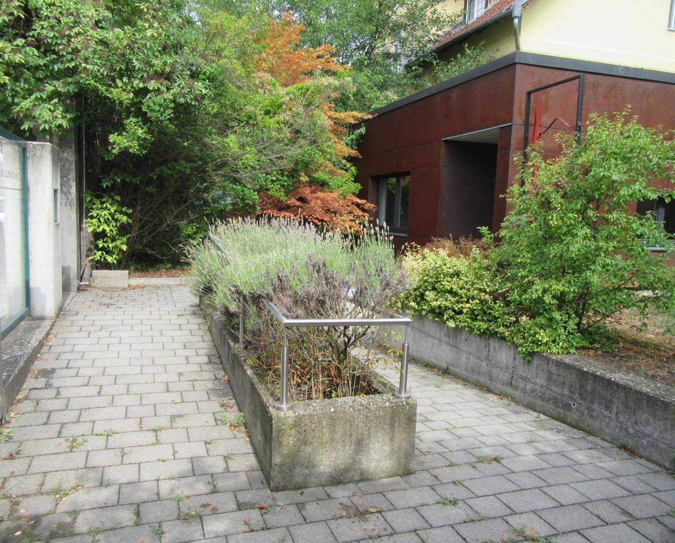 Gepflasterter Zugangsbereich mit einer Rampe, einem bepflanzten Hochbeet und üppigem Grün.