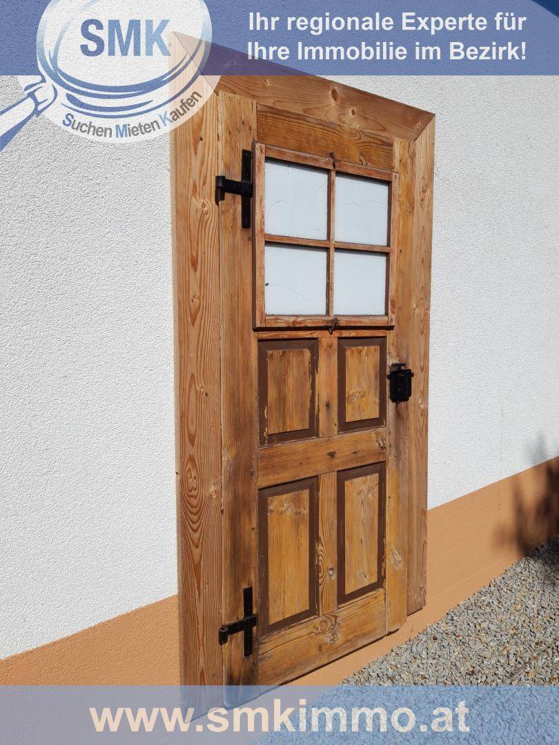 Rustikale Holztür mit kleinem Fenster und schwarzen Beschlägen in weißer Wand.