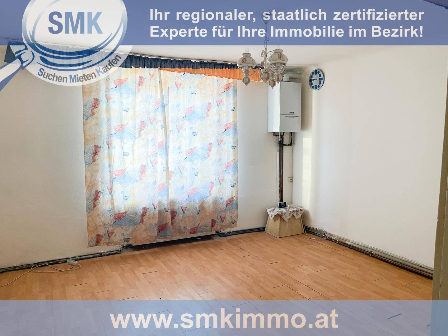 Leeres Zimmer mit altem Parkettboden, Gastherme an der Wand und Vorhängen am Fenster.