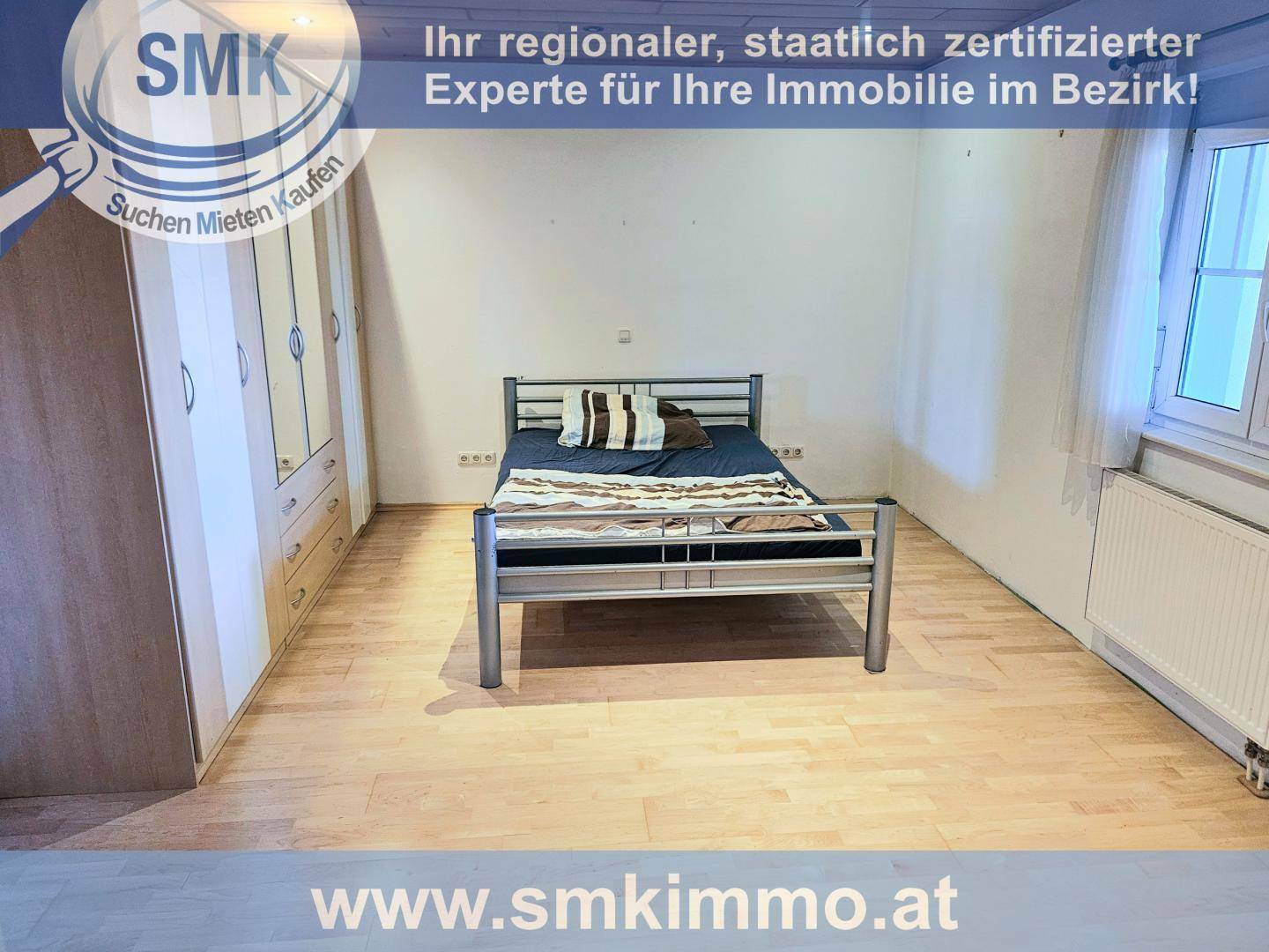 Schlafzimmer mit Holzboden, Metallbett und großem Kleiderschrank, hell durch Fenster.