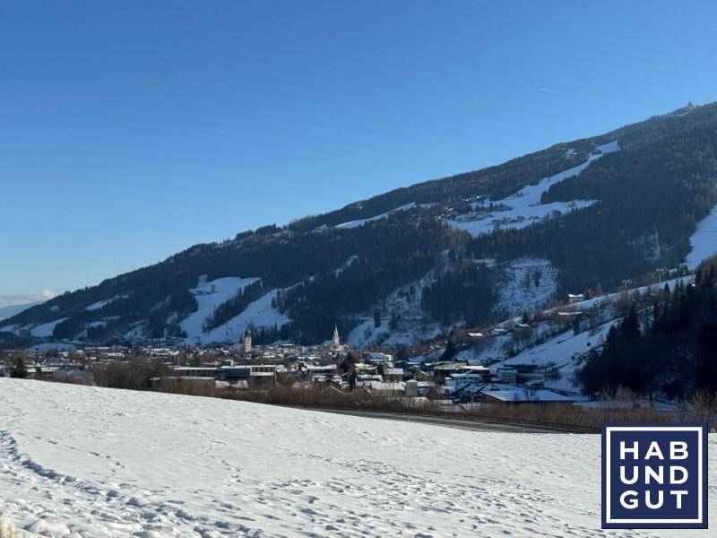 Winterlandschaft mit schneebedeckten Feldern, einem Dorf und Bergen unter klarem blauem Himmel.